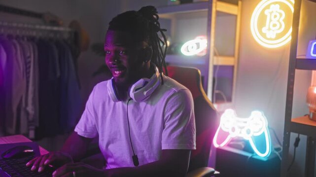 Man with headset sitting in gaming chair typing on keyboard and giving thumbs up in studio lit by neon controller and bitcoin sign; fun streaming joy.