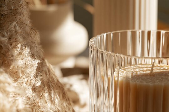 Close-up of a fluted glass holding a textured cream candle with soft pampas grass and a blurred vase in the background, illuminated by warm sunlight