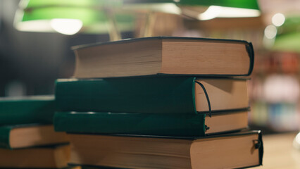 Stack of books rests on a desk in a quiet university library with warm lighting and a green lamp, surrounded by study materials and wooden bookshelves in an empty academic reading interior. © DC Studio