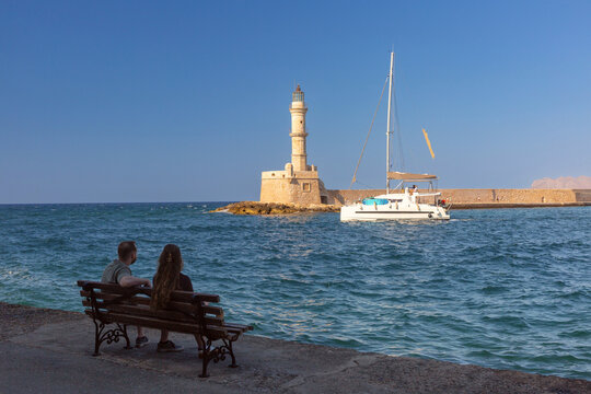 Couple sitting on a waterfront bench overlooking a sailboat and the historic lighthouse at the Old Venetian Harbor in Chania, Crete, Greece.