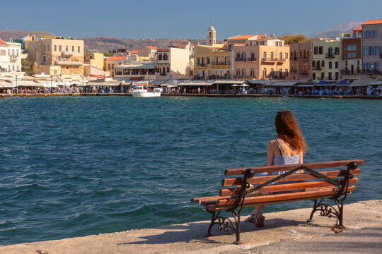 Woman sitting on a waterfront bench overlooking the colorful historic buildings and sea of the Old Venetian Harbor in Chania, Crete, Greece.