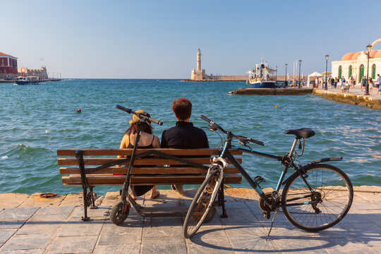 Couple sitting on a waterfront bench beside a bicycle and electric scooter overlooking the Venetian Harbor and historic lighthouse in Chania, Crete, Greece.