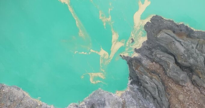 FPV aerial view of the turquoise crater lake of Ijen Volcano with rugged rocky cliffs and mineral deposits in East Java, Indonesia.