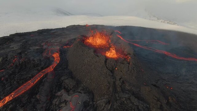 FPV aerial view of glowing lava flowing from an active volcanic crater across dark solidified lava fields with snow-covered mountains in the background in Iceland.