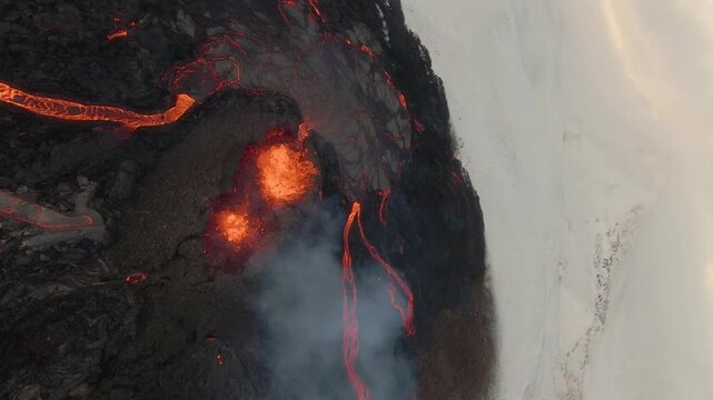 FPV aerial view of glowing lava flowing from a volcanic crater across dark solidified rock with rising smoke in Iceland.