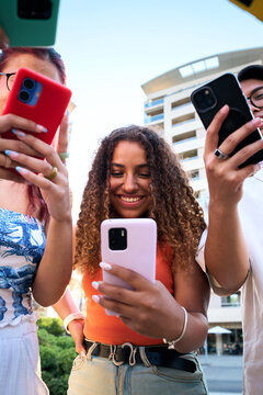 Vertical Diverse group of young friends using smartphones together outdoors smiling while checking messages and social media. Low-angle view digital communication friendship and modern youth lifestyle