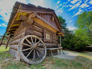 Cabin with wheel under blue sky. The wooden cabin features a wagon wheel on the ground. It is surrounded by trees in a rural area during daylight. © Анна Терелюк