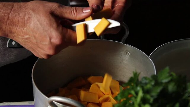 Hands carefully slice bright orange butternut squash cubes into a large metal pot, preparing for a hearty meal. hands slicing pumpkin  halloween