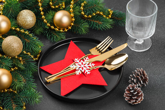 Holiday table setting with cutlery arranged in red star shaped holder decorated with white snowflake on black plate, wine glass and Christmas tree branch with godlen balls on dark textured background