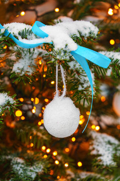 Snow covered Christmas tree branches decorated with white ornaments and a blue ribbon on glowing fairy lights colorful bokeh background. Cozy winter holiday atmosphere.
