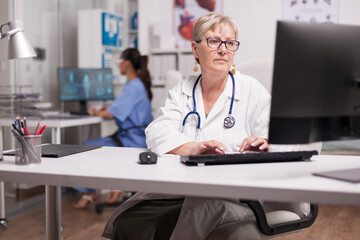 Senior female doctor types at her workstation while a nurse works behind her, combining focused...