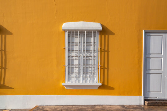 Colonial window with bright yellow wall frames a white barred window. White window casts crisp shadow on vivid surface. Window grate shows vertical and horizontal bars. Simple design evokes quiet. Pue