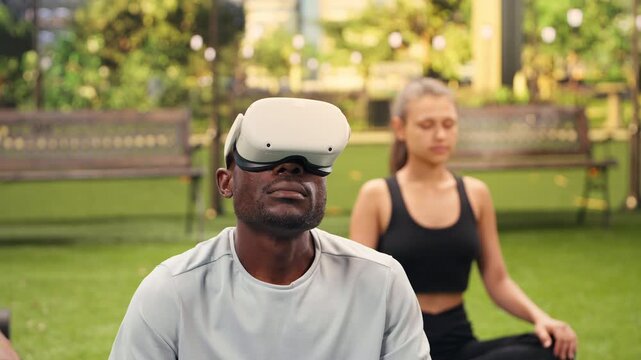 Man wearing VR headset sits cross-legged on yoga mat in city park, beginning outdoor meditation with virtual reality in lotus position alongside participants during a Pilates fitness session.