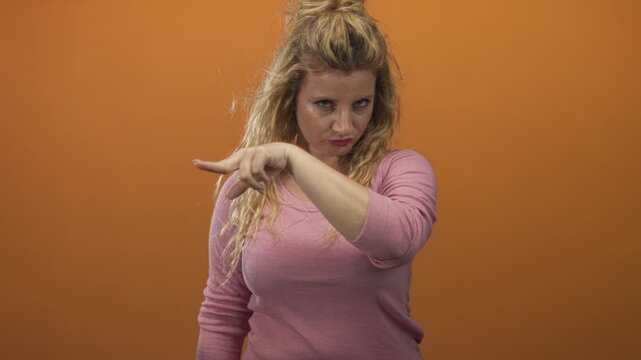 Woman with blonde hair points finger at camera in studio with orange backdrop and focused stare wearing pink top; confidence empowerment challenge.