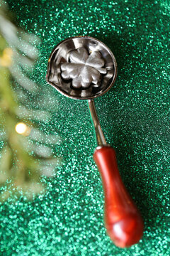 metal melting spoon holding silver clover shapes on a sparkling green background with evergreen branches, illustrating New Year fortune telling, good luck and traditional European holiday rituals 