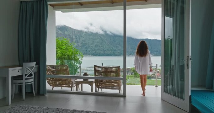 Woman in white shirt walking to resort hotel balcony, enjoy serene morning view of Lake Batur and surrounding mountains in Bali, Indonesia, vacation, travel, peaceful silence. Back view tracking shot