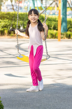 A young girl in bright clothing standing on a yellow swing at a sunny park.