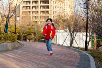 Full shot of a happy school girl with pigtails in a red puffer jacket and jeans carrying a pink backpack, walking along a paved garden path and smiling with blurred background. © Li Liu
