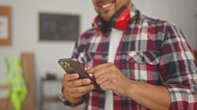 Man tapping smartphone with finger, wearing earmuffs and plaid shirt in building; productivity concentration.