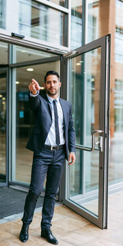 A man in a suit and tie standing at an open glass door, pointing forward with his index finger. He appears to be directing someone or indicating a path