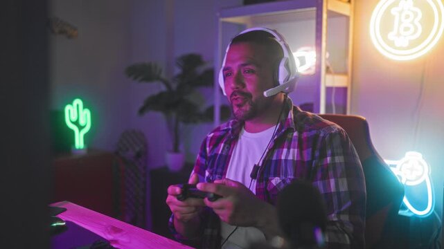Man with headset holds a game controller, leaning forward at his desk and gripping the controller with visible hands in a gaming studio lit by neon signs and rgb lights; excitement celebration.