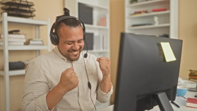 Man wearing headset and microphone smiling and fist pumping at a computer monitor in an office building while seated and leaning forward; work success joy.