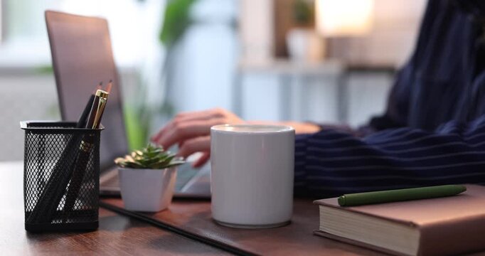 Woman working with laptop and taking mug of coffee at wooden table indoors, closeup