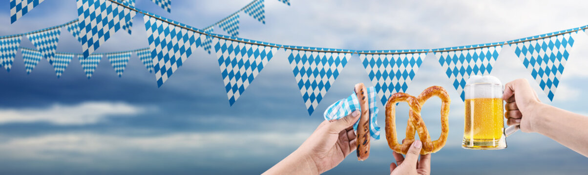 Bavarian Oktoberfest celebration banner with blue and white diamond pattern flags, pretzel, sausage, and a mug of beer against a cloudy sky background. Bavarian Oktoberfest Festival