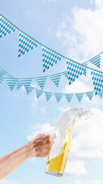 A person holds a large glass beer mug, spilling and splashing beer against a background of bright blue sky with white clouds and traditional Bavarian checkered pennants. Bavarian Oktoberfest Festival