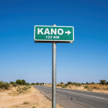 Green road sign pointing to Kano 125 km distance on a dusty highway under a clear blue sky