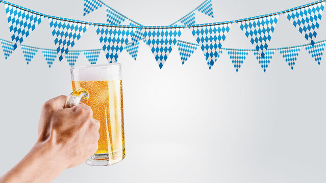 Hand holding a large beer stein with foaming beer against a background of Bavarian blue and white diamond pattern pennant banners for Oktoberfest celebration. Bavarian Oktoberfest Festival
