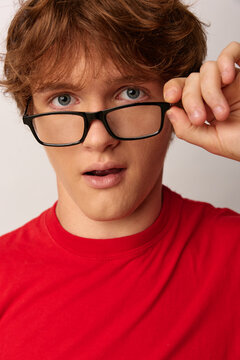 Extreme close up of a young man with red hair and wide blue eyes looking over his glasses in disbelief. He wears a red t-shirt on a gray background. Concept for surprise and doubt.