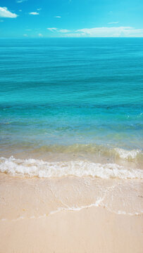 Crystal clear turquoise water, gentle white waves lapping onto smooth sandy beach, vibrant blue sky, idyllic tropical seascape backdrop. Nature, natural, or quotes background