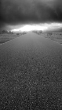 Low angle black and white shot of an empty asphalt road leading into the distance under dramatic storm clouds with light on the horizon. Quotes background