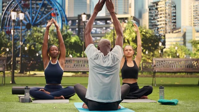 Yoga instructor demonstrates a raised arms stretch as women follow on fitness mats in city park, supporting mobility and guided wellness exercise.