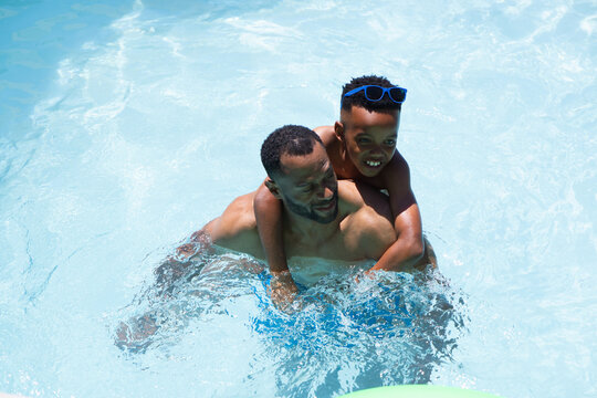 African American father and son splashing in sunlit pool with blue sunglasses, trunks, green float