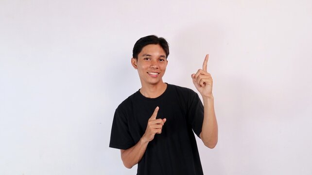 Cheerful young Asian man smiling warmly while pointing one index finger up and the other to the side against a white studio background.