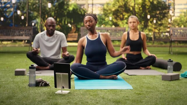 Woman instructor does a side stretch in front of a tablet with participants, following a digital yoga tutorial together on mats in a public park.
