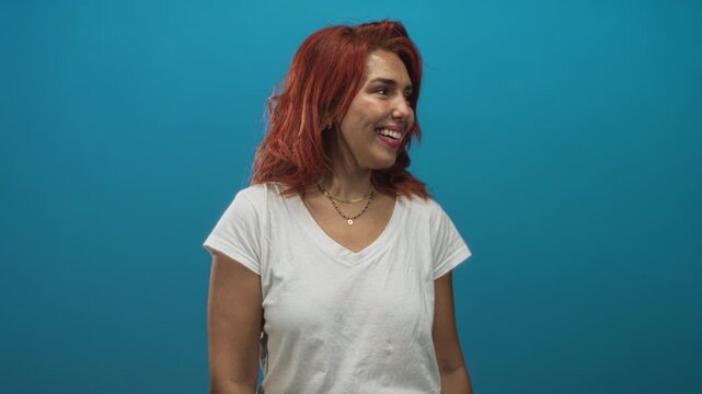 Woman smiling with visible face and red hair, wearing a white t shirt against a teal studio wall; joy confidence openness.