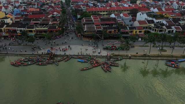 Busy Quay With Tourists Boarding Small Wooden Boats In Hoi An Shoreline Crowded With Pedestrians And Vendors, Colorful Hulls Tied In Clusters, Lively Evening Mood And Heritage Storefronts