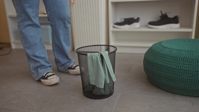 Woman drapes a green shirt over a mesh wastebasket rim while standing in a fitting room building; indecision shopping choice.