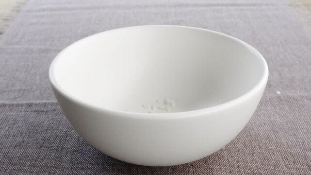White round rice grains pouring and filling white bowl to half on burlap fabric, 4K
