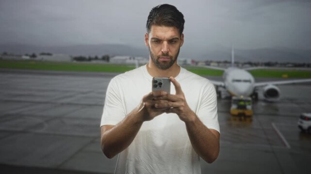 Man with beard holding smartphone and tapping screen with both hands at airport near airplane in white t shirt; focus travel planning.