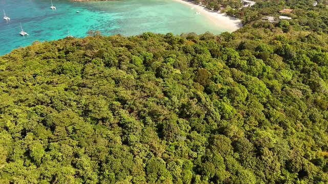 Aerial flight over lush tropical forest canopy toward a turquoise bay with anchored boats in Racha Island Phuket Thailand