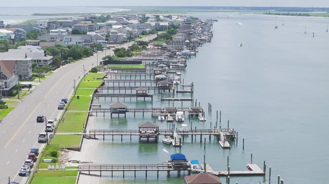 Aerial view shows coastal town with docks and boats. Water reflects bright sunlight and calm surface. Road runs parallel with parked cars and green lawns. Boats rest quietly under blue tarps