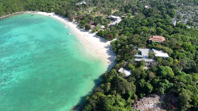 Aerial view of a curved white sand beach with turquoise shallow water bordered by jungle and small villas in Racha Island Phuket Thailand