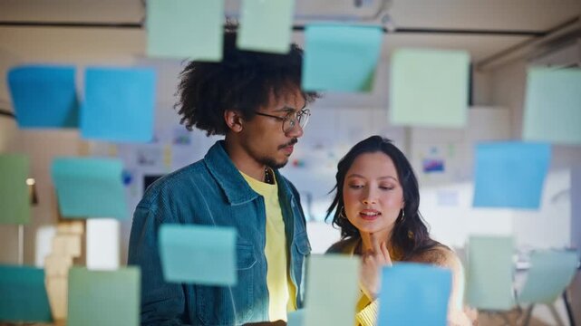Coworkers arranging sticky notes glass wall. Multiethnic pair planning ideas.