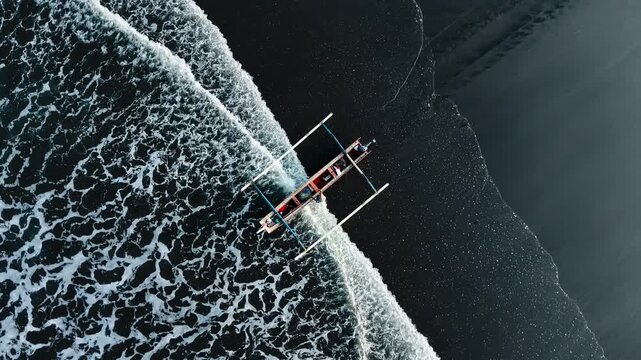 Top down drone view of traditional outrigger boat jukung surrounded by white foamy ocean waves on black sand beach. Dramatic action seascape finished vibrant color for cinematic film