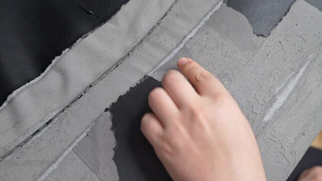 Close view of a hand testing a flaking gray surface on a black artificial leather seat, revealing poor material quality and a concept of consumer disappointment with cheap furnishing.