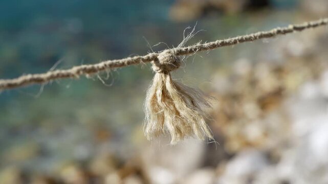 A natural jute rope with a frayed knot and loose fibers sways slightly in the wind, set against a soft blurred pebble shore and turquoise water at the seaside.